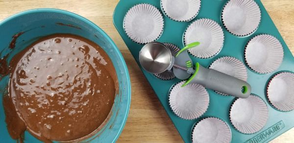 a blue bowl with chocolate cake mix batter in it next to a blue muffin tin with cupcake liners in it and a scoop on top, all on a brown table