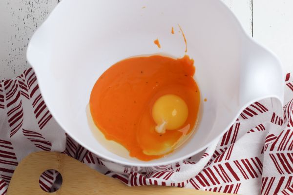 buffalo sauce and an egg in a white mixing bowl on a red and white cloth next to a wooden spoon