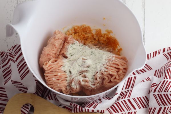 ground chicken, ranch seasoning, panko bread crumbs, buffalo sauce and an egg in a white mixing bowl on a red and white cloth next to a wooden spoon