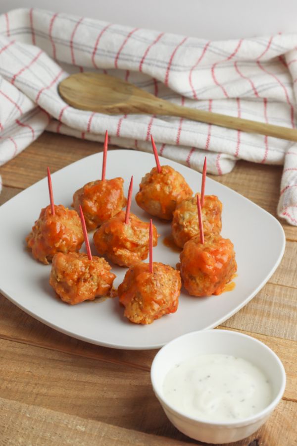 buffalo chicken meatballs with toothpicks in them on a white plate on a brown table next to a white bowl of sauce with a wooden spoon on a red and white cloth in the background