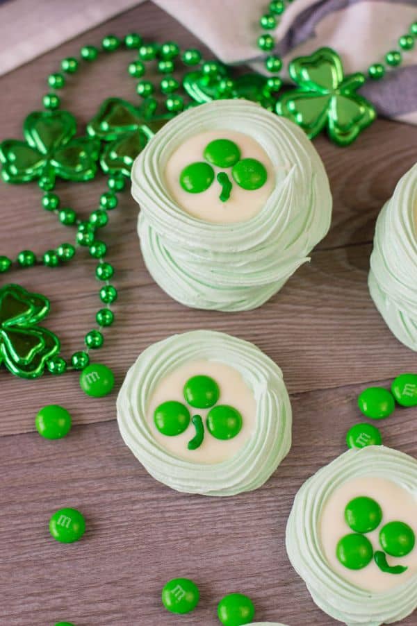 green meringue cookies topped with melted white candy melts, three green m&ms, and a stripe of green frosting to look like a clover, all on a brown table next to green beads with shamrocks on them 