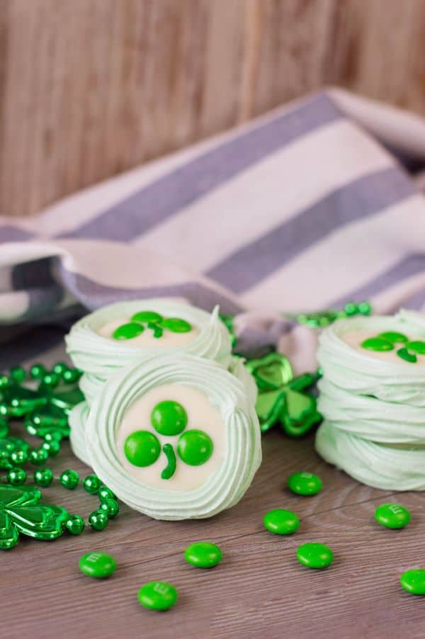 green meringue cookies topped with melted white candy melts, three green m&ms, and a stripe of green frosting to look like a clover, all on a brown table next to green beads with shamrocks on them and more green m&ms with a white and blue striped cloth in the background