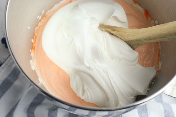 orange jello powder, whipped topping and Greek yogurt in a metal mixing bowl with a wooden spoon on a striped cloth