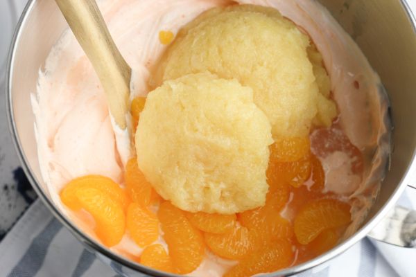 orange jello powder, whipped topping, Greek yogurt, mandarin oranges, and pineapple in a metal mixing bowl with a wooden spoon on a striped cloth