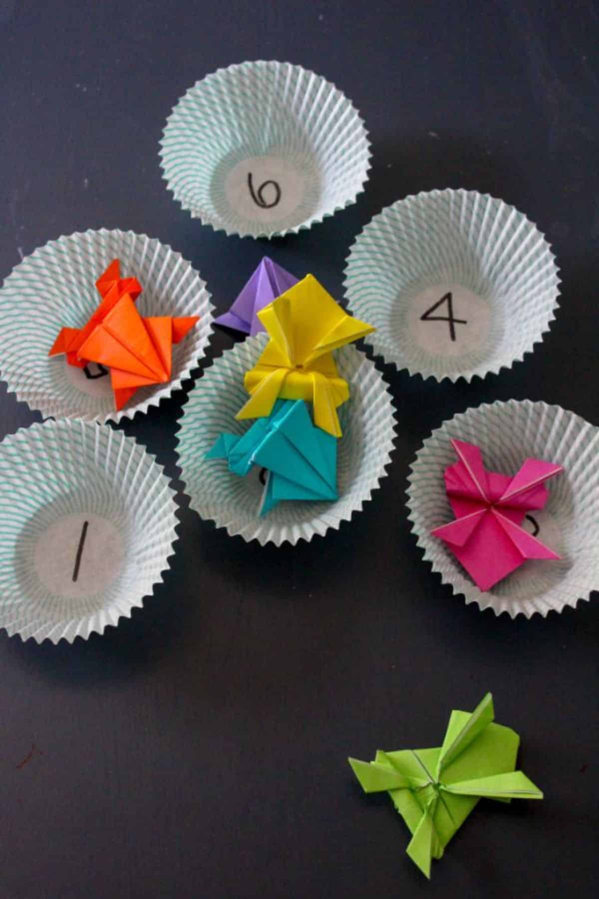 Origami colored paper jumping frogs in  cupcake liners with numbers written on them. 