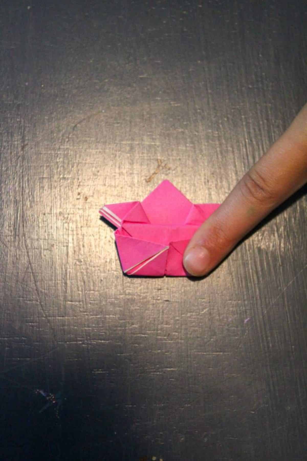 a finger touching pink origami paper folded to look like a frog on a black background.