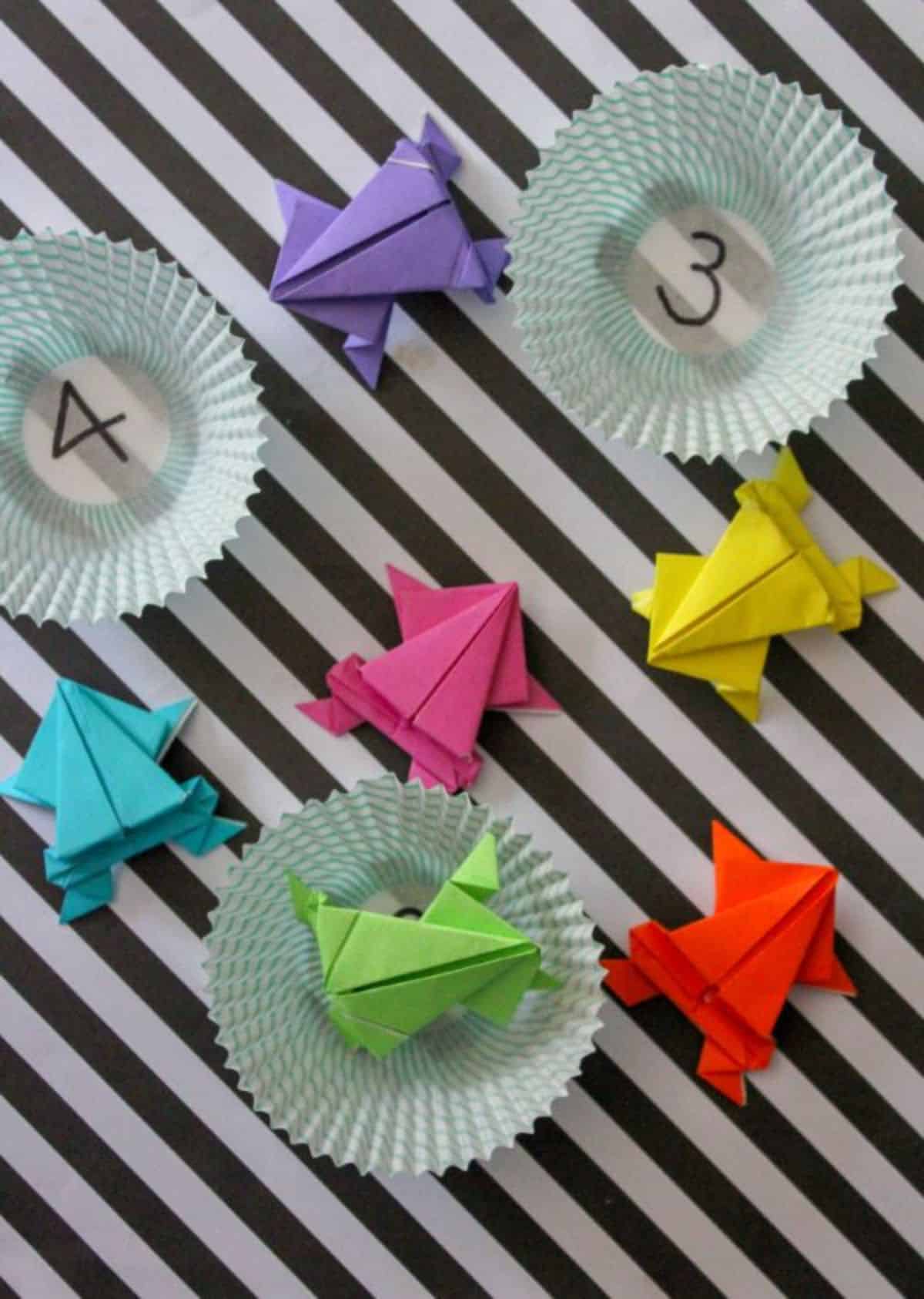 Origami frogs and cupcake liners with numbers written on them on a black and white striped background.