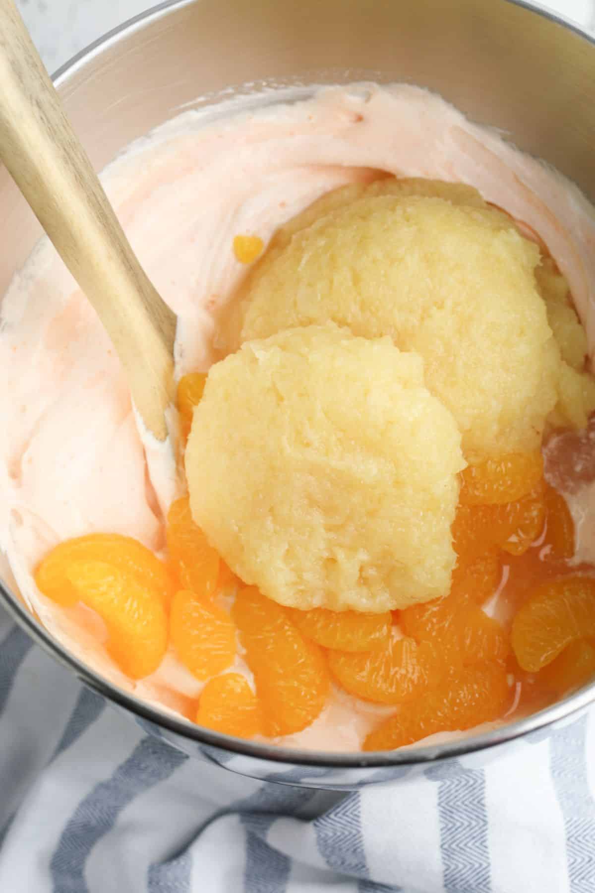 orange jello powder, whipped topping, Greek yogurt, mandarin oranges, and crushed pineapple in a metal mixing bowl with wooden spoon.