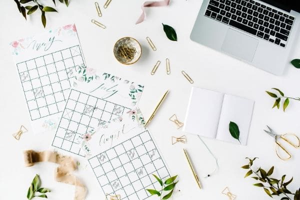 a desk with a laptop, calendar pages, paper clips, pen, and notebook