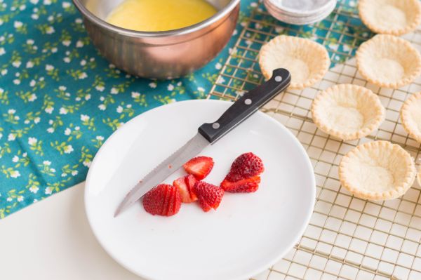 halved strawberries and a knife on a white plate next to tart shells on a wire rack and a bowl of custard on a blue cloth with flowers on it