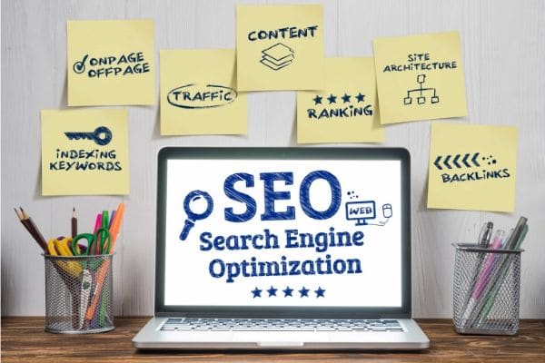 a laptop on a desk with text on the screen reading SEO Search Engine Optimization,. There's also two pencil cups on the desk and sticky notes on the wall behind it with text reading indexing keywords, on page off page, traffic, content, ranking, site architecture and backlinks