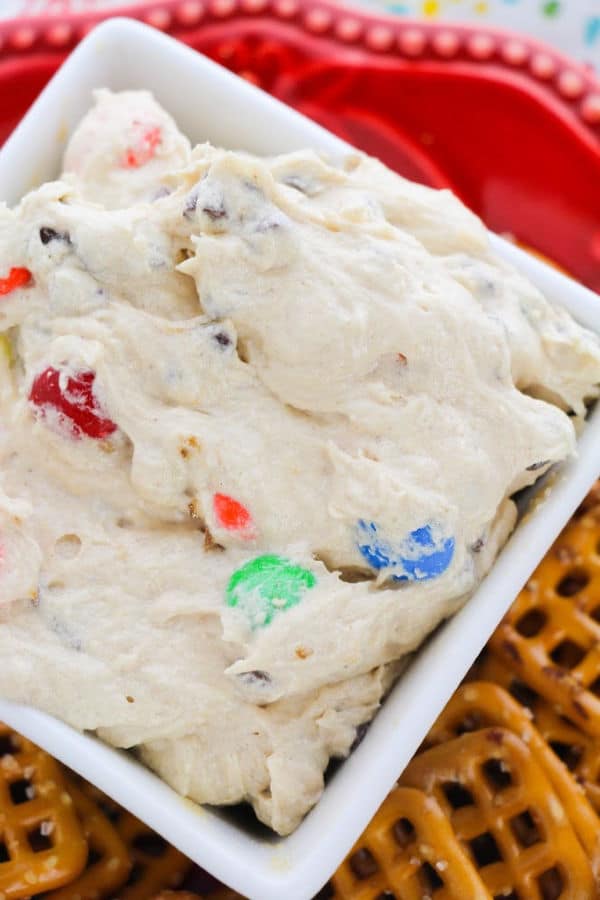 close up view of Cream Cheese Monster Cookie Dough Dip in a white dish next to pretzels on a red plate on a multi-colored cloth