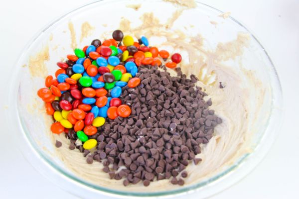 m & m's, chocolate chips, and the rest of the Cream Cheese Monster Cookie Dough in a glass bowl on a white table