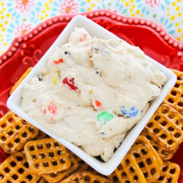 Cream Cheese Monster Cookie Dough Dip in a white dish next to pretzels on a red plate on a multi-colored cloth