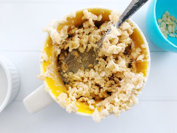 a spoon being used to stir Microwave Rice Krispie Treat for One in a mug on a white wood table next to a blue bowl with a few rice crispie cereal pieces in it