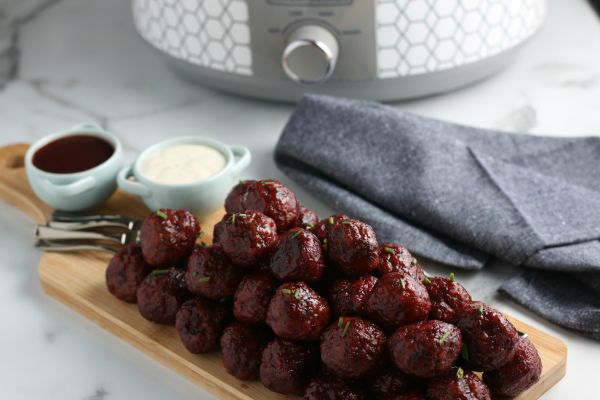 easy crockpot grape jelly BBQ meatballs next to forks and  sauce in  bowls on a wooden board with a gray cloth and slow cooker in the background