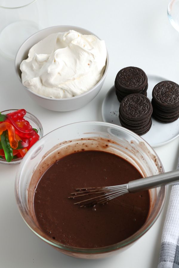 bowls of pudding, whipped cream and gummy worms, with oreos on a plate