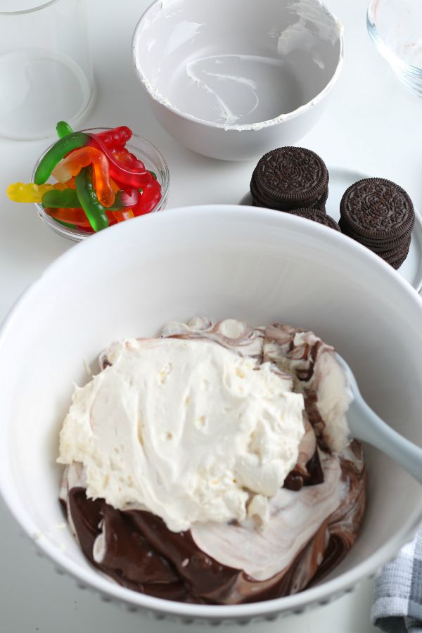 whipped cream and chocolate pudding being combined in a bowl, next to a bowl of gummy worms and a plate of oreo cookies