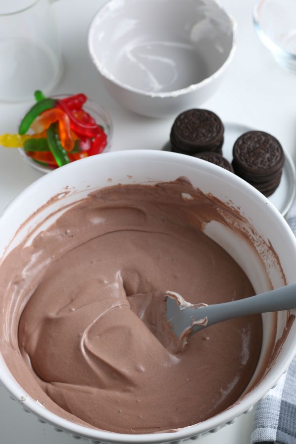 whipped cream and chocolate pudding combined in a bowl, next to a bowl of gummy worms and a plate of oreo cookies