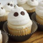 Ghost cupcakes with white frosting shaped like friendly spirits, each topped with two chocolate chips for eyes, are arranged in paper liners on a wooden surface.