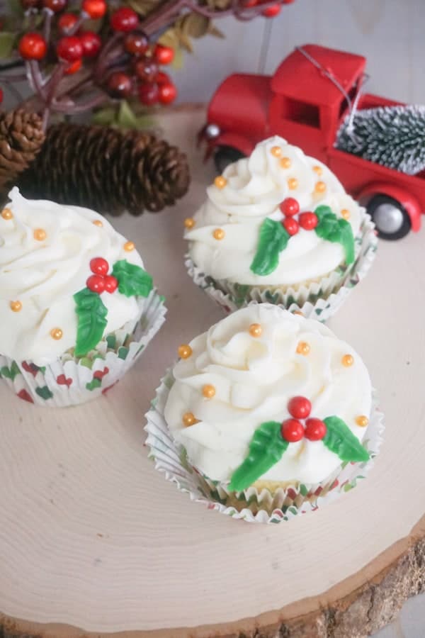 three Christmas holly cupcakes on a log with pincecones and a red truck in the background