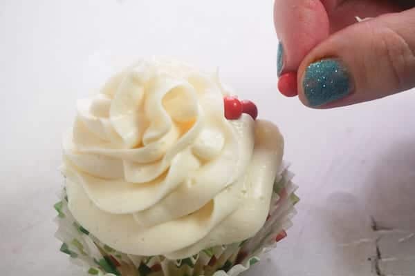 a hand putting red candy balls on a frosted cupcake