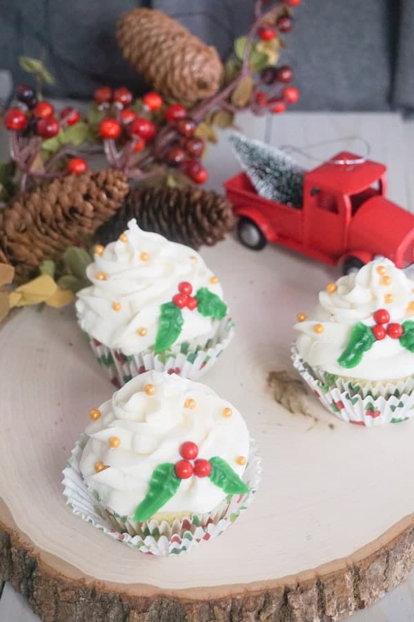 three Christmas holly cupcakes on a log with pincecones and a red truck in the background