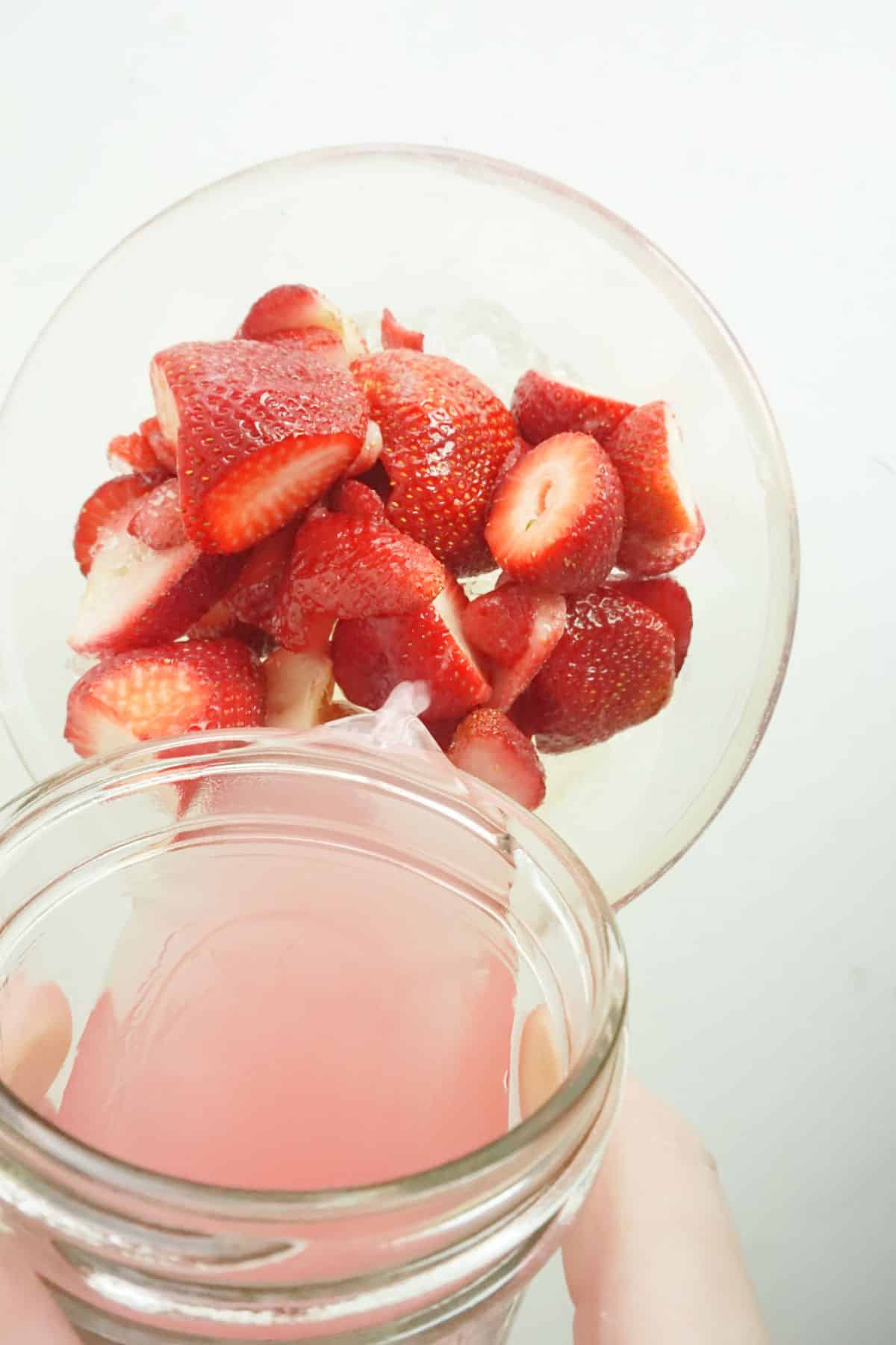 strawberry lemonade is being poured from a glass jar into a glass bowl of strawberries.