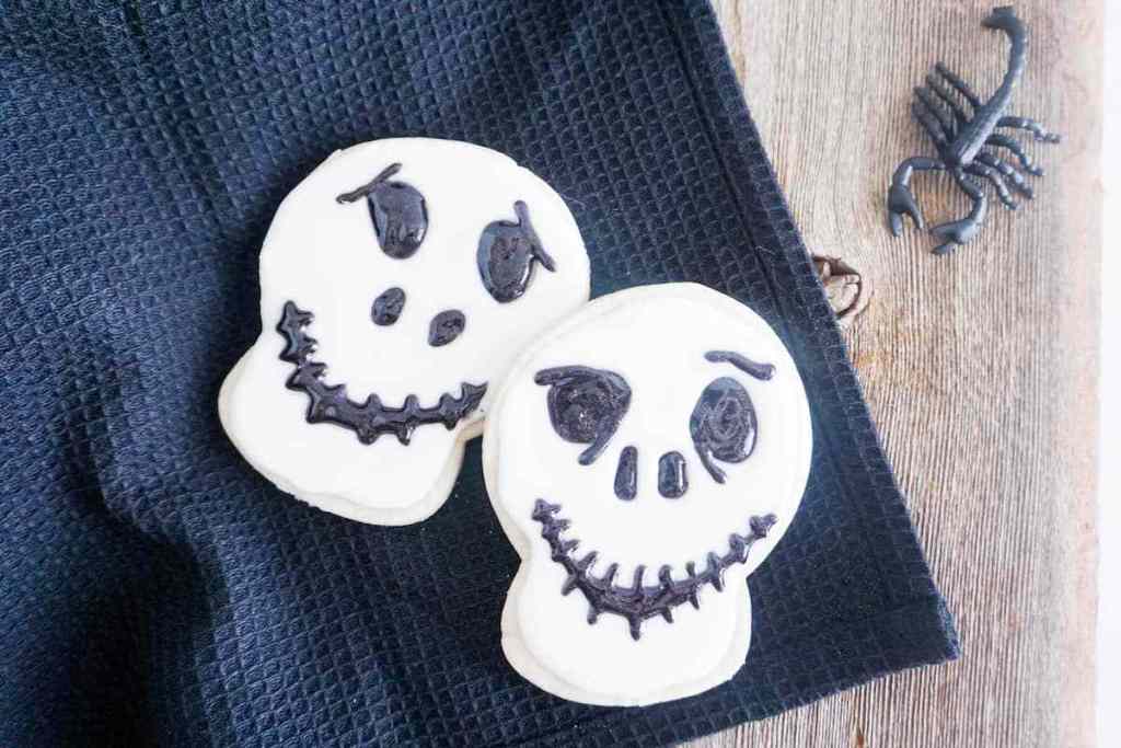 two cookies decorated with white frosting and black gel to look like Jack Skellington on a black linen on a wood table with a fake scorpion  next to it