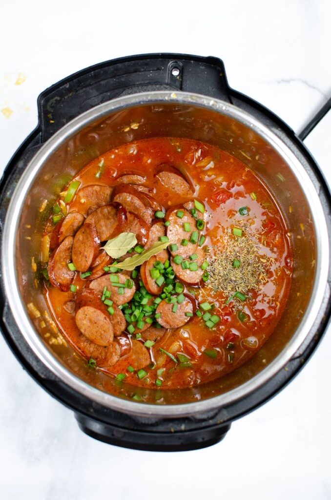 Chopped sausages, green onions and bay leaves on top of other ingredients in an instant pot.