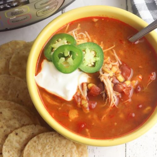 Mexican tortilla soup topped with sour cream and jalapenos in a yellow bowl next to tortilla chips and an instant pot.