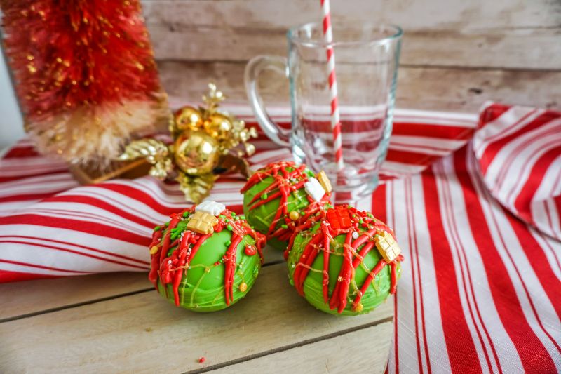 Christmas Hot Cocoa Bombs on wooden serving tray next to a red and white striped cloth and a glass mug with a straw in it