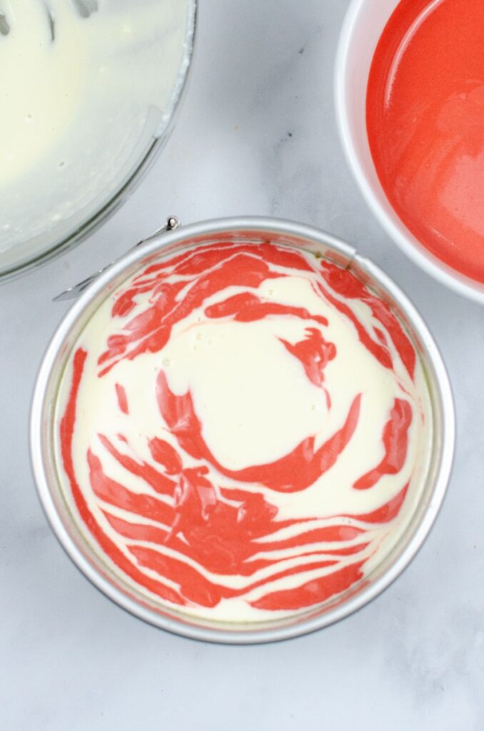 Red velvet swirl cheesecake in a tin next to pots of cheesecake mixture.