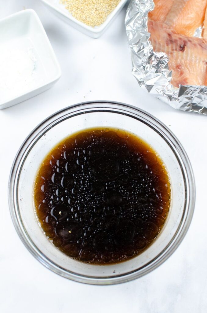 Soy sauce mixture in a glass bowl next to baked salmon in aluminium foil and white bowls.