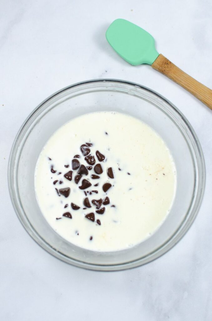 Milk and chocolate chips in a glass bowl with a spatula at the side.
