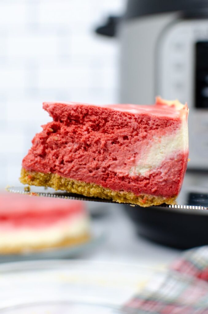 Slice of red velvet cheesecake on a knife in front of an instant pot.