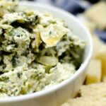 A white bowl of spinach artichoke dip in another white bowl of bread cubes.
