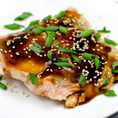 Baked salmon in Teriyaki Sauce on a white plate garnished with sesame seeds and green onions.