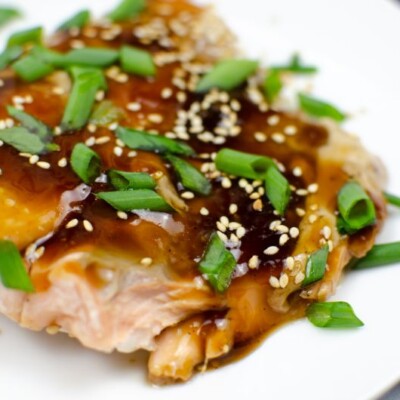 Slice of baked salmon in Teriyaki sauce covered with sesame seeds and green onions on a white plate.