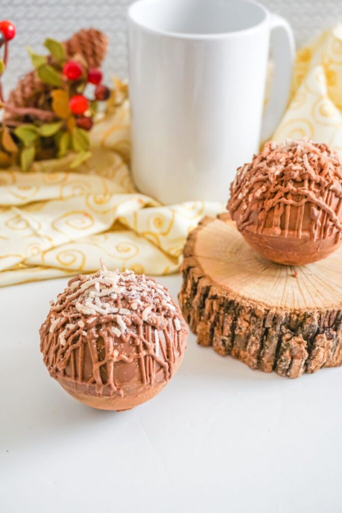 Mounds hot cocoa bombs on a table and wooden log with a white mug on a yellow cloth in the background
