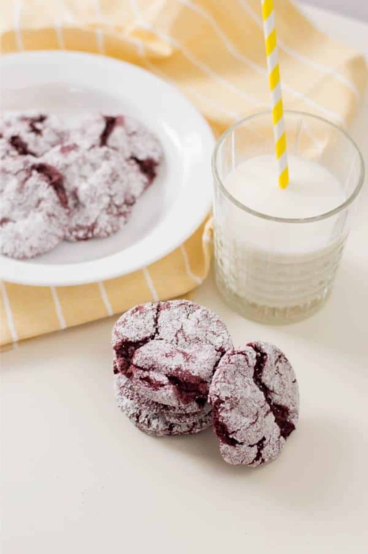 red velvet cookies stacked on a white table with more cookies on a white plate in the background on a yellow cloth next to a glass of milk.