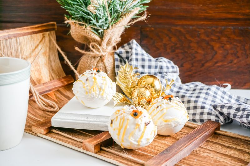 Snowman Hot Cocoa Bombs on wooden platter with a mini Christmas tree and a black and white checkered cloth in the background