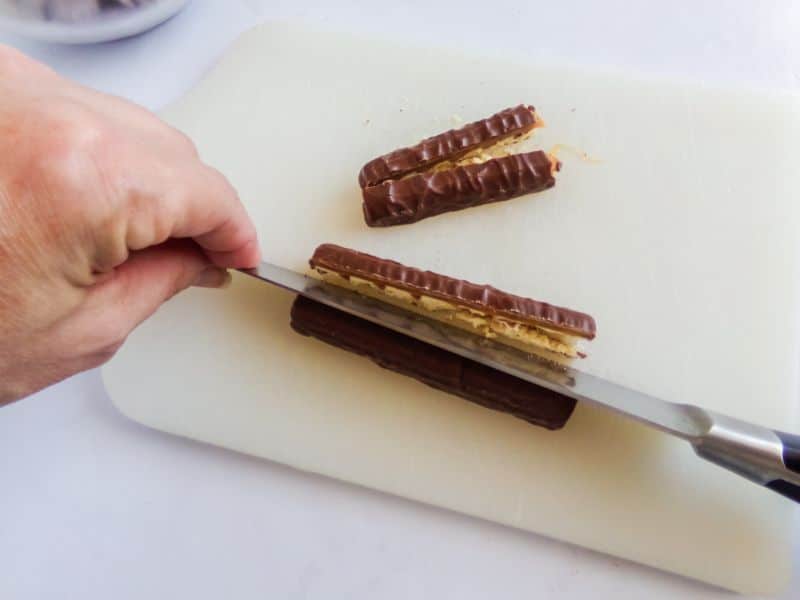 a hand using a knife to cut twix candy bars in half on a white cutting board