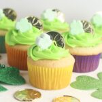 Shamrock cupcakes adorned with green frosting and festive shamrock decorations are topped with playful coins. They're artfully displayed alongside green heart and shamrock cutouts on a pristine white surface, creating a charming scene.