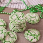 A stack of vibrant green crinkle cookies with powdered sugar rests on a wooden surface, surrounded by scattered shamrock beads.