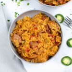 A bowl of rice with sliced sausage, bell peppers, and spices reminiscent of Instant Pot Jambalaya. Fork and sliced jalapeños are nearby on a white surface.