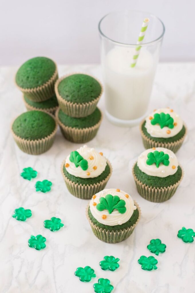 Homemade Green Velvet Cupcakes decorated for St. Patrick's Day next to shamrocks and a glass of milk on a white background