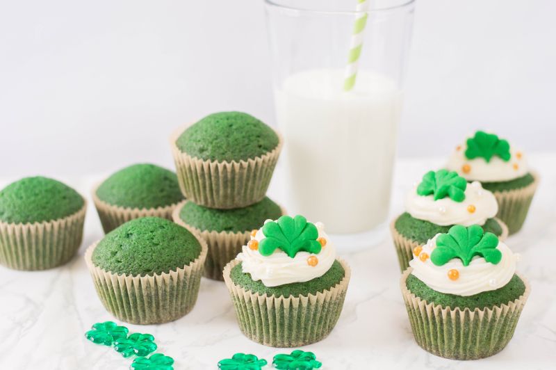 Homemade Green Velvet Cupcakes decorated for St. Patrick's Day next to shamrocks and a glass of milk on a white background