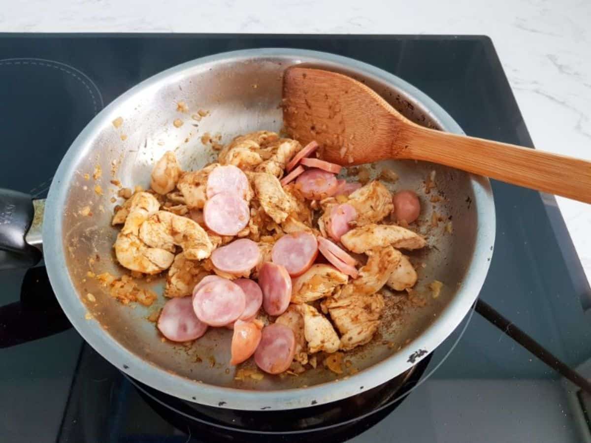 smoked sausage, chicken pieces and onions cooking in oil in a skillet, wooden spatula on the side.