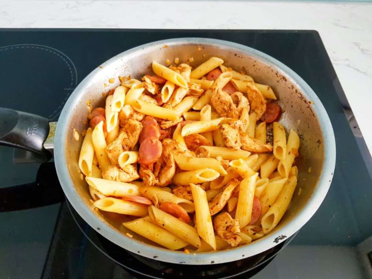 cooked pasta and water, smoked sausage, chicken pieces and onions cooking in a skillet.